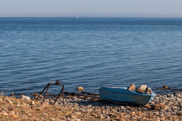 Fototapeta na wymiar Fishing boat on the shore of the blue Lake Baikal. Pressed down by large stones. Near the iron device. Away ship. On the horizon of the mountain.