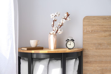 Vase with cotton branches, clock and cup on table in bedroom