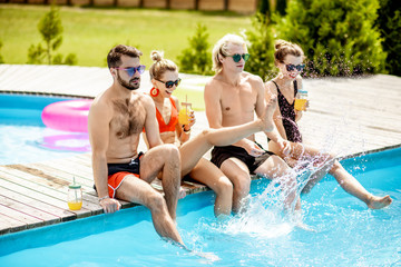 Group of happy friends sitting together on the water poolside, talking and having fun splashing water, enjoying the summer time