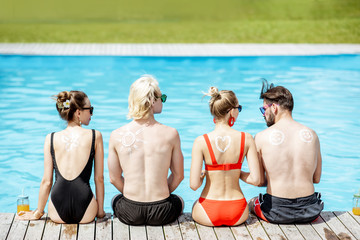 Friends with sun cream painted shapes on the shoulders sitting together on the poolside. Concept of a skin protection from a sun, rear view