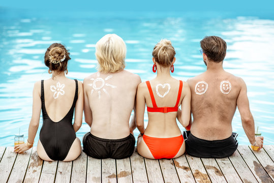Friends with sun cream painted shapes on the shoulders sitting together on the poolside. Concept of a skin protection from a sun, rear view