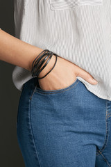 Cropped top and bottom photo of a woman with a bracelet on her arm. The model on a gray background is dressed in a light striped shirt and jeans. You can see a multi-layer bracelet made from leather a