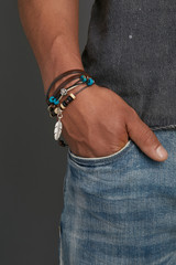 Cropped top and bottom photo of a man with a bracelet on his arm. The model on a gray background is dressed in a gray shirt and jeans. You can see a multi-layer bracelet made from leather and laces wi