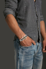 Cropped from top and bottom view shot of a man with a bracelet on his arm. The fashion model on the gray background is dressed in a gray shirt and jeans. You can see a multi-layer bracelet made of lea