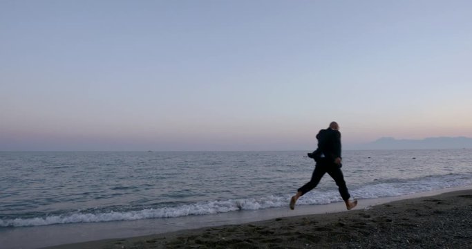 Happy adult bald man with glasses and business suit, quietly walking barefoot along the calm evening sea.