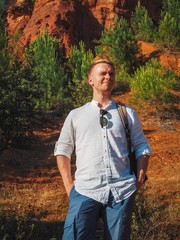 A man in a white shirt and a backpack stands on the slope of the Ochre trail in Roussillon in France, orange stones