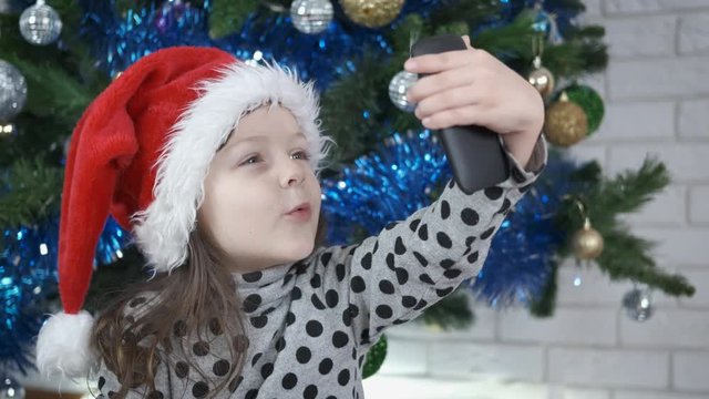 Child plays with grimaces. A cute little girl is photographed on a smartphone in a santa claus hat.