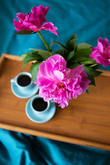 Beautiful pink peonies in a glass vase and two cups of coffee are standing on a wooden tray in the bed.