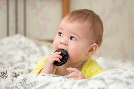 Little European Baby Girl Boy Puts In His Mouth The Cap From The Camera Lens. A Novice Photographer For 7 Months Lies On The Bed And Sucks The Lens Cover. A Child Pulls In The Mouth Lying On His Stoma