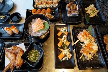 Variety of food with fried chicken wings, grilled bacon wrapped golden needle mushroom, Crab stick salad, sushi and sashimi on dark background in restaurant, food of hotel buffet, ready to eat