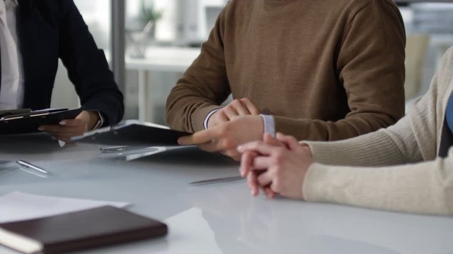 Medium body shot of unrecognizable man and woman sitting at table in notary office, professional lady giving them documents on clipboards, and couple starting to read, tracing lines with fingers