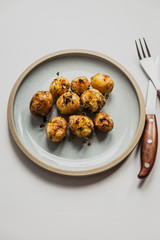 A vertical photo of baked baby potatoes with salt, pepper, olive oil and thyme. Natural light, white background, blue plate, fork and knife.