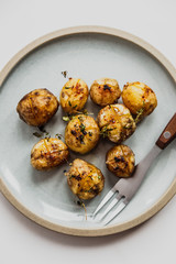 A top view vertical photo of baked baby potatoes with salt, pepper, olive oil and thyme. Natural light, white background, blue plate, fork.