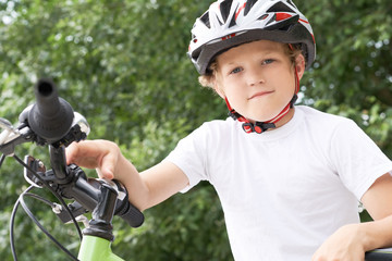 Small Caucasian boy in protective helmet stands leaning on the bike posing for the camera. Teenager ready to ride bicycle in park on summer day. Weekend activity.