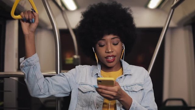Portrait of young african american woman wearing i with headphones listening to music, sing and funny dancing in public transport. He holds the handrail.