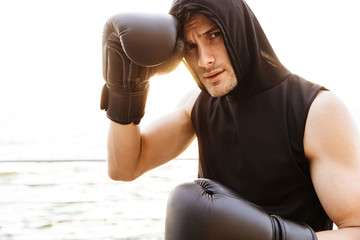 Photo of young man wearing hoodie working out in black boxing gloves on wooden pier at seaside