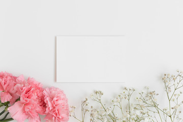 Top view of a white card mockup with gypsophila and a bouquet of pink carnations on a white table.