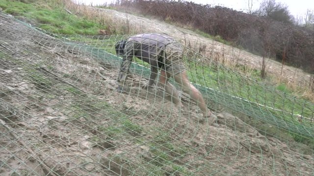 An obstacle course racer crawls under a cargo net up a hill