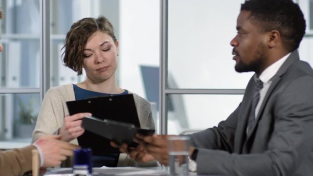 Tracking waist-up shot of Afro-American broker consulting young Caucasian client couple in company office, giving them draft copies of home insurance policy and explaining terms and conditions