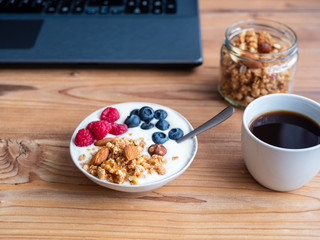Joghurt mit Müsli und frischen Himbeeren und Heidelbeeren im Büro, Kaffee, Frühstück am Arbeitsplatz