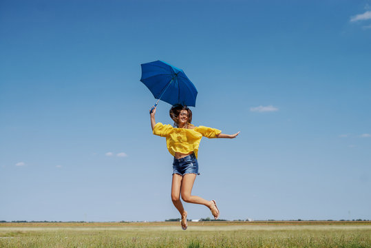 Cheerful Caucasian teenage girl in yellow blouse, denim shorts and with eyeglasses jumping outdoor with blue umbrella in hand.