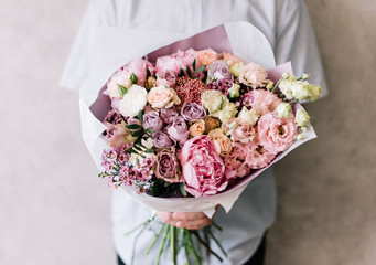 very nice young man holding a beautiful blossoming flower bouquet of fresh peony, roses, eustoma,carnations, in tender pink colors on the grey wall background