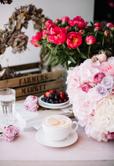 Delicious morning cup of cappuccino coffee with fresh summer cherries, blueberries and strawberries on a plate, big pink hydrangea, peony and roses flower bouquet  with coral peonies on the background