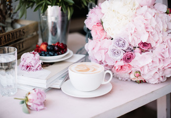 Delicious morning cup of cappuccino coffee with fresh summer cherries, blueberries and strawberries on a plate, big pink hydrangea, peony and roses flower bouquet  with coral peonies on the background