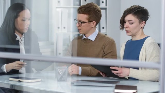 Hidden camera handheld shot through window of Asian female law practitioner consulting casually dressed Caucasian client couple in her office and giving them paperwork on clipboards to read and sign