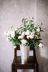 Big bucket of fresh white peonies standing on a wooden rustic bar stool on the grey wall background