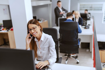 Gorgeous Caucasian businesswoman in formal wear using computer and talking on the phone. In...