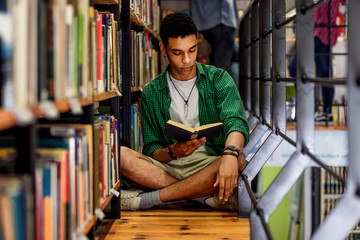 Young male student study in the library reading book while sitting near bookshelf.