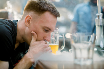 Close-up shot of a stylish adult guy smoking hookah indoor of a cafe, sitting at the table together with his friends.