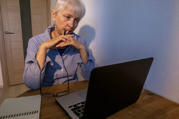 An elderly woman in a blue-white shirt and blue T-shirt looks sadly at the monitor. Nervously nibbles the handle. Emotions: depression, frustration, fright