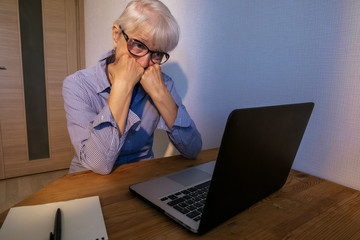 An elderly caucasian woman in a blue-white shirt and blue T-shirt sadly looks at the monitor. Operated hands on fists. Emotions: depression, frustration, fright
