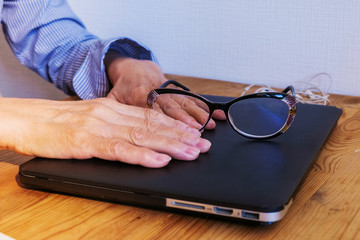 Hands of an elderly woman holding glasses on the lid of a closed laptop. Closeup