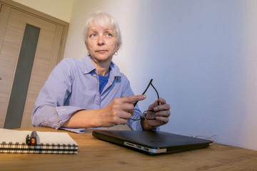 Pensive caucasian elderly woman sits at a laptop and holds glasses