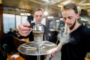 Two young boys preparing shisha by placing the charcoals on top. One Man lighting hookah on bar background.