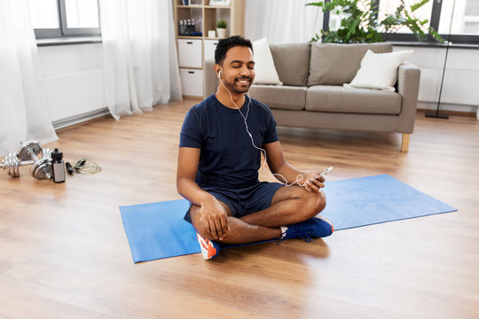 fitness, meditation and healthy lifestyle concept - indian man in earphones listening to music on smartphone and meditating in lotus pose at home