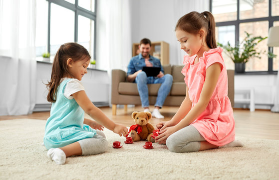 childhood, leisure and family concept - little sisters playing tea party game with toy crockery and teddy bear at home