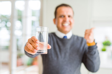Middle age man drinking glass of water at home pointing and showing with thumb up to the side with happy face smiling