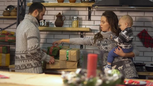 Pretty young woman with a baby in her arms and bearded man in warm sweater make out gifts presents boxes. Happy family celebrating Christmas together
