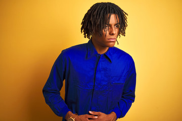 Afro worker man with dreadlocks wearing mechanic uniform over isolated yellow background with hand on stomach because nausea, painful disease feeling unwell. Ache concept.