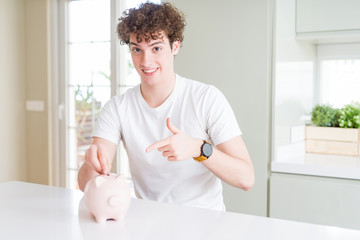 Young man investing money using piggy bank at home very happy pointing with hand and finger