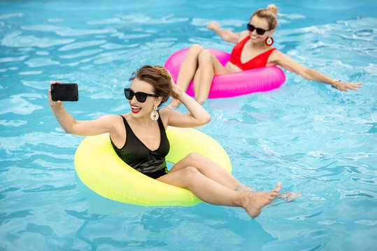 Girlfriends making selfie photo with phone while swimming on inflatable rings in the water pool outdoors during the summertime