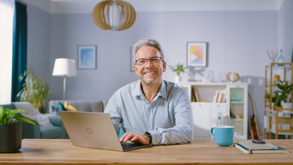 Handsome Middle Aged Man Uses Laptop Computer and Smiles on Camera. He is in the Cozy and Stylish Living Room. Happy Successful Man Works on Computer from Home.