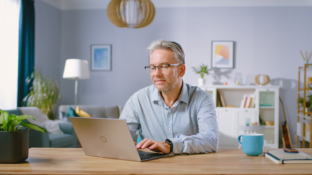 Handsome Middle Aged Man Uses Laptop Computer While Sitting at His Desk in the Cozy and Stylish Living Room. Happy Successful Man Works on Computer from Home.