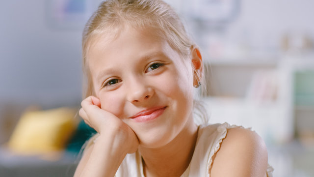 Portrait of a Cute Little Blonde Girl Smiling, She's Sitting at the Desk of a Cozy Living Room. Child Smils on Camera.