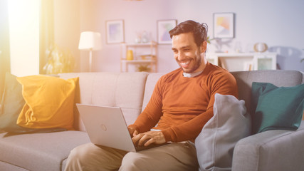 Cheerful Young Man Sitting on a Sofa Holds Laptop on His Lap, Browses Through the Internet, Social Networks, Does e-Shopping. Man at Home Using Laptop while Sitting on a Couch. Warm Sun Light.