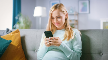 Beautiful Happy Pregnant Woman Sitting on a Couch Browses Through Internet on Smartphone. Future Mom Does Internet Shopping on Smartphone from Home.